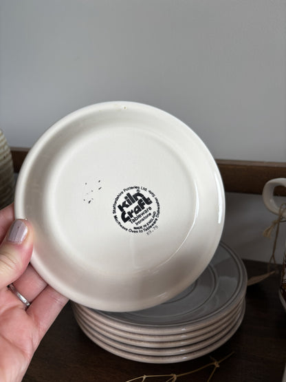 A hand holds a round off-white ceramic plate showing a black printed circular logo in the center. A stack of similar plates sits on a dark wooden surface below, with a neutral indoor background.