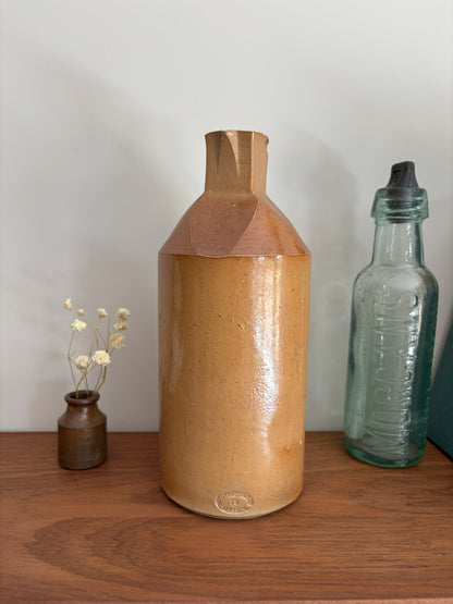 A two tone brown vintage ink bottle with a textured, glossy tan-brown glaze and a geometric faceted neck, displayed on a wooden shelf next to a small vase of dried flowers and a vintage glass bottle.