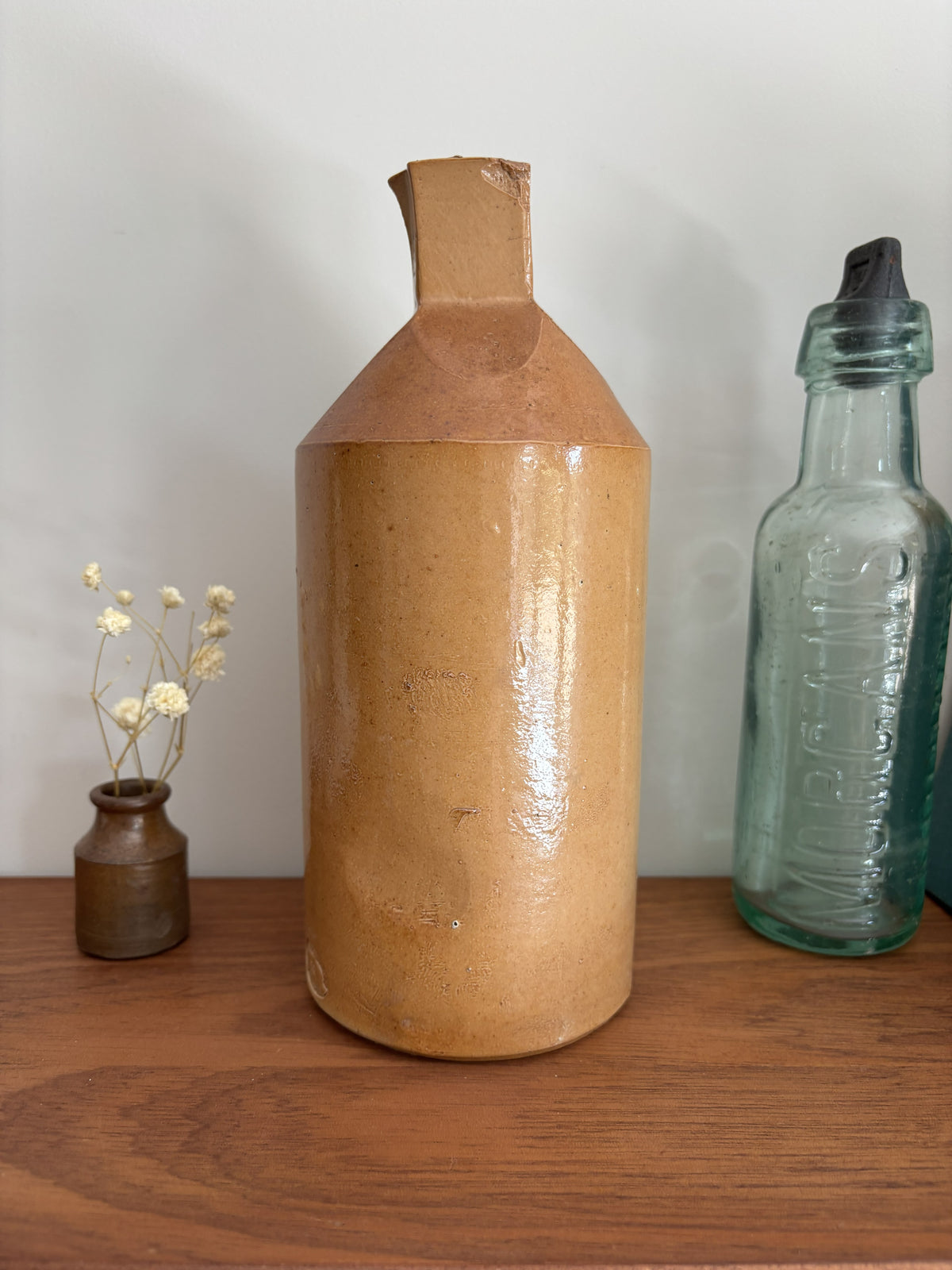 A two tone brown vintage ink bottle with a textured, glossy tan-brown glaze and a geometric faceted neck, displayed on a wooden shelf next to a small vase of dried flowers and a vintage glass bottle.