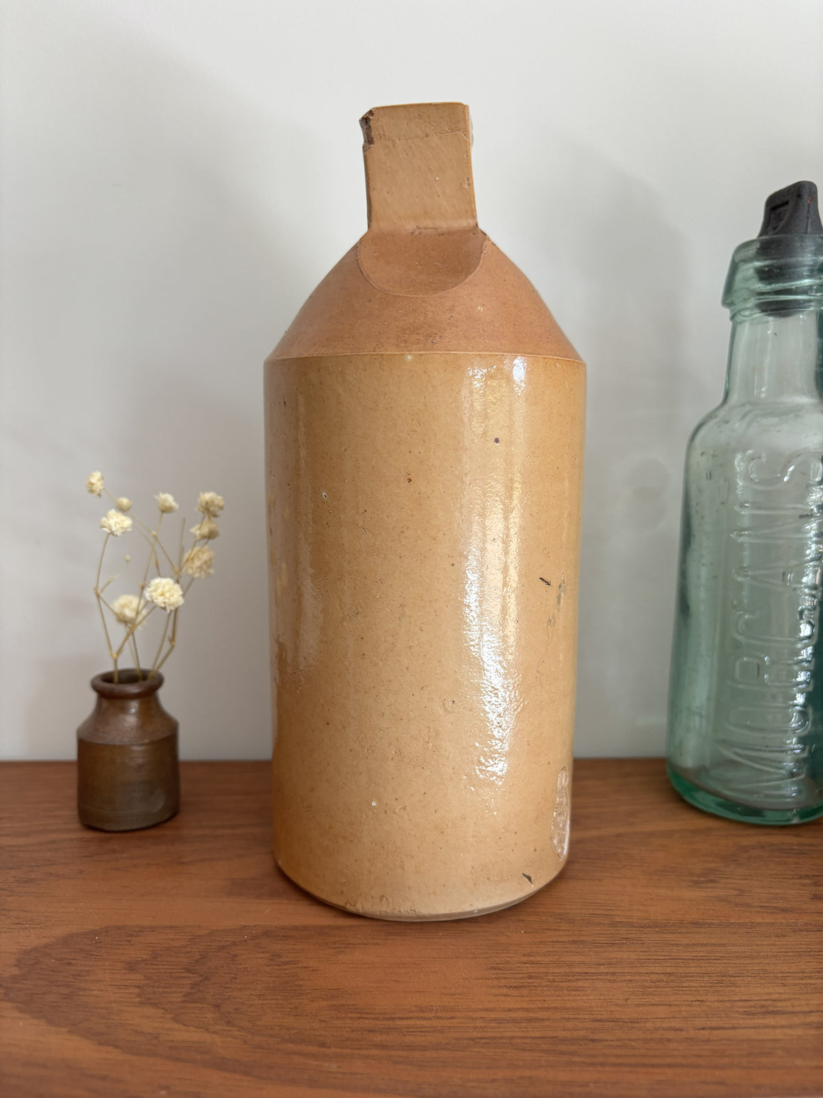 A two tone brown vintage ink bottle with a textured, glossy tan-brown glaze and a geometric faceted neck, displayed on a wooden shelf next to a small vase of dried flowers and a vintage glass bottle.