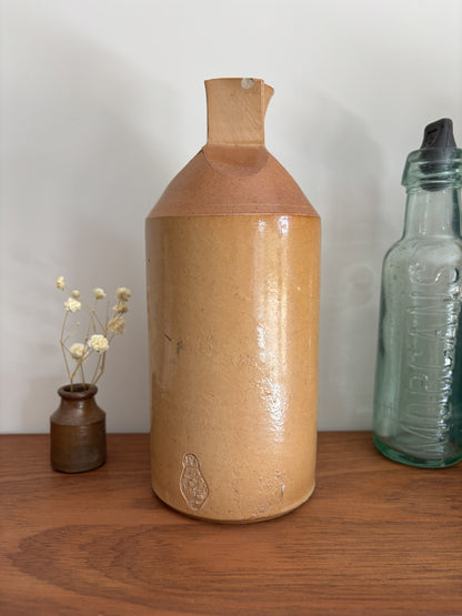 A two tone brown vintage ink bottle with a textured, glossy tan-brown glaze and a geometric faceted neck, displayed on a wooden shelf next to a small vase of dried flowers and a vintage glass bottle.