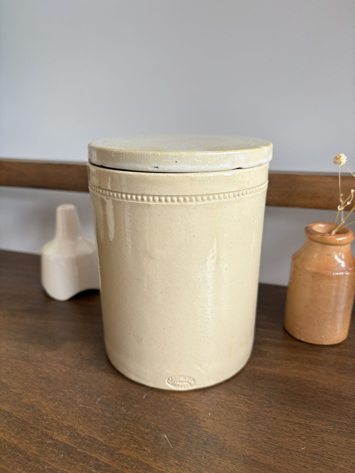 A cream-colored glazed ceramic canister with a matching lid, displayed on a wooden surface next to two small decorative vases.