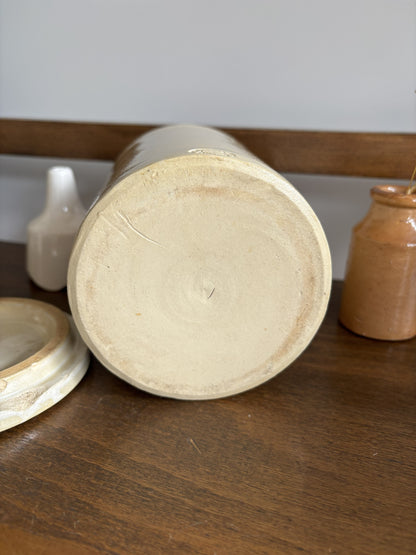 The base of a ceramic jar with a light beige stoneware finish, shown lying on its end on a dark wooden table. Its matching lid rests to the left, and two small rustic clay bottles are visible in the background.