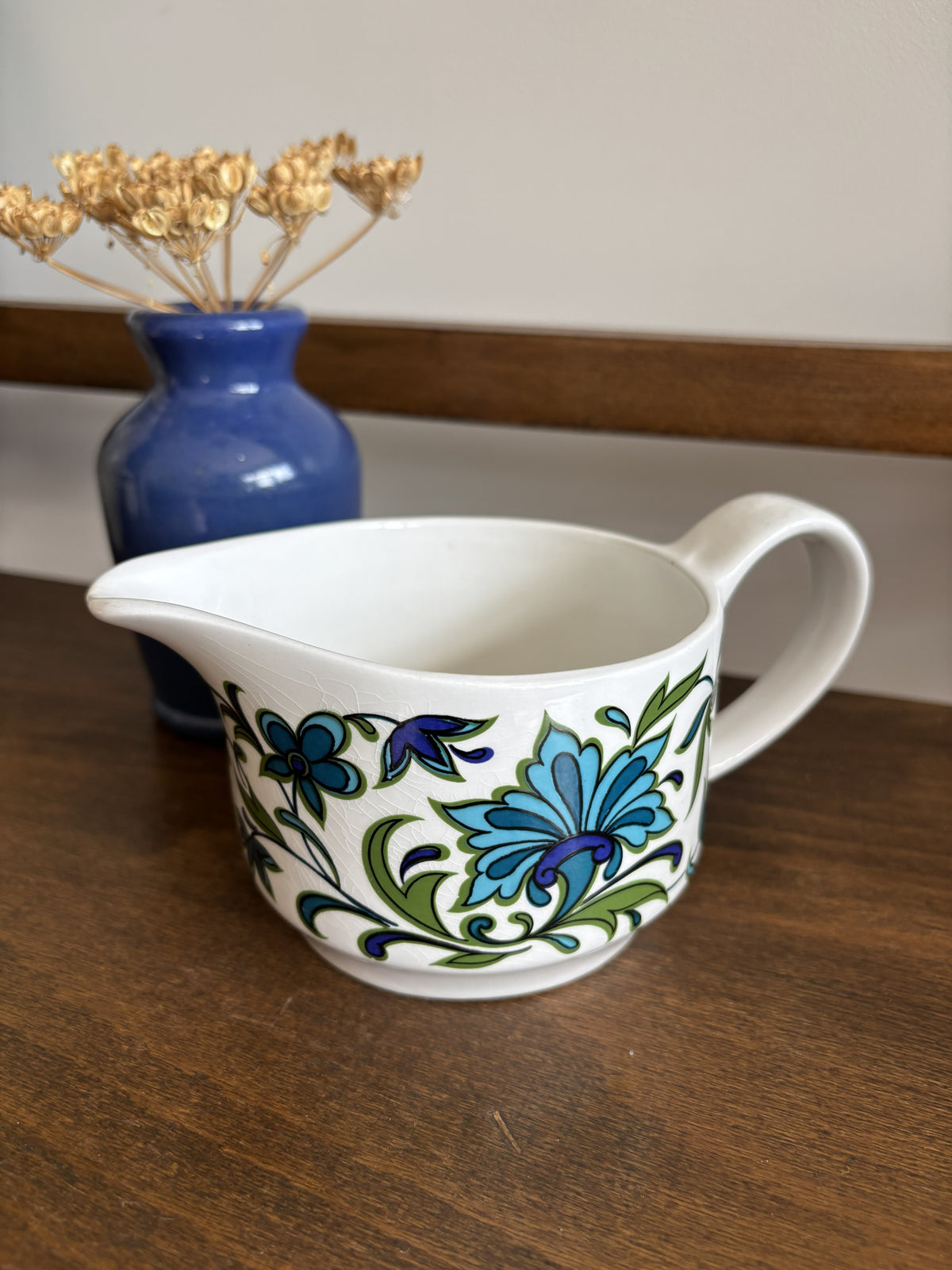 A white ceramic jug with a blue and green floral pattern, placed on a wooden table next to a blue vase with dried flowers.