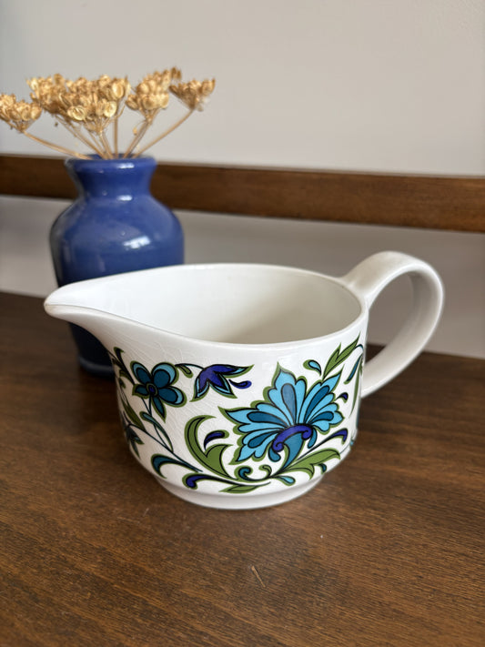 A white ceramic jug with a blue and green floral pattern, placed on a wooden table next to a blue vase with dried flowers.