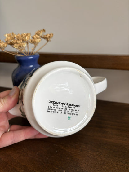 A hand holds the bottom of a white ceramic jug, showing the printed 'Midwinter' brand mark and manufacturing details. The background features a wooden table and a blue vase with dried flowers.