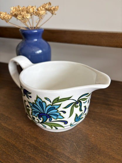 A white ceramic jug with a blue and green floral pattern, placed on a wooden table with a blue vase of dried flowers in the background.