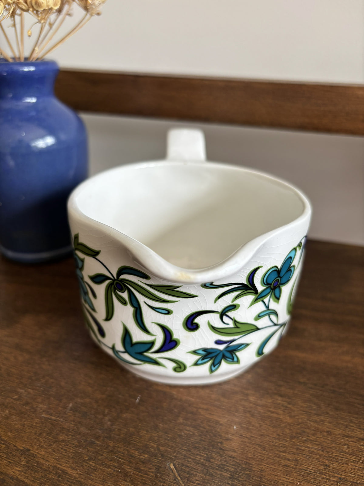A white ceramic milk jug with a blue and green floral pattern, sitting on a wooden table next to a blue vase.