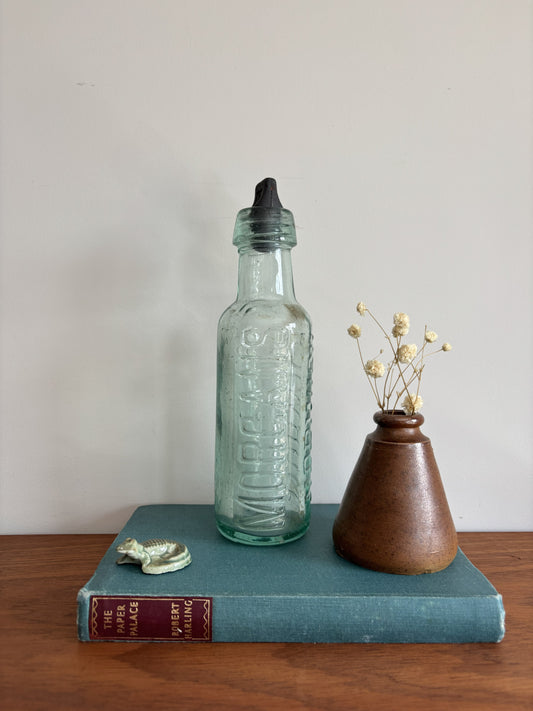 A vintage glass bottle with a black stopper, displayed on a teal book next to a small brown ceramic vase with dried flowers.  The words Morgans and Norwich are embossed on the sides. 