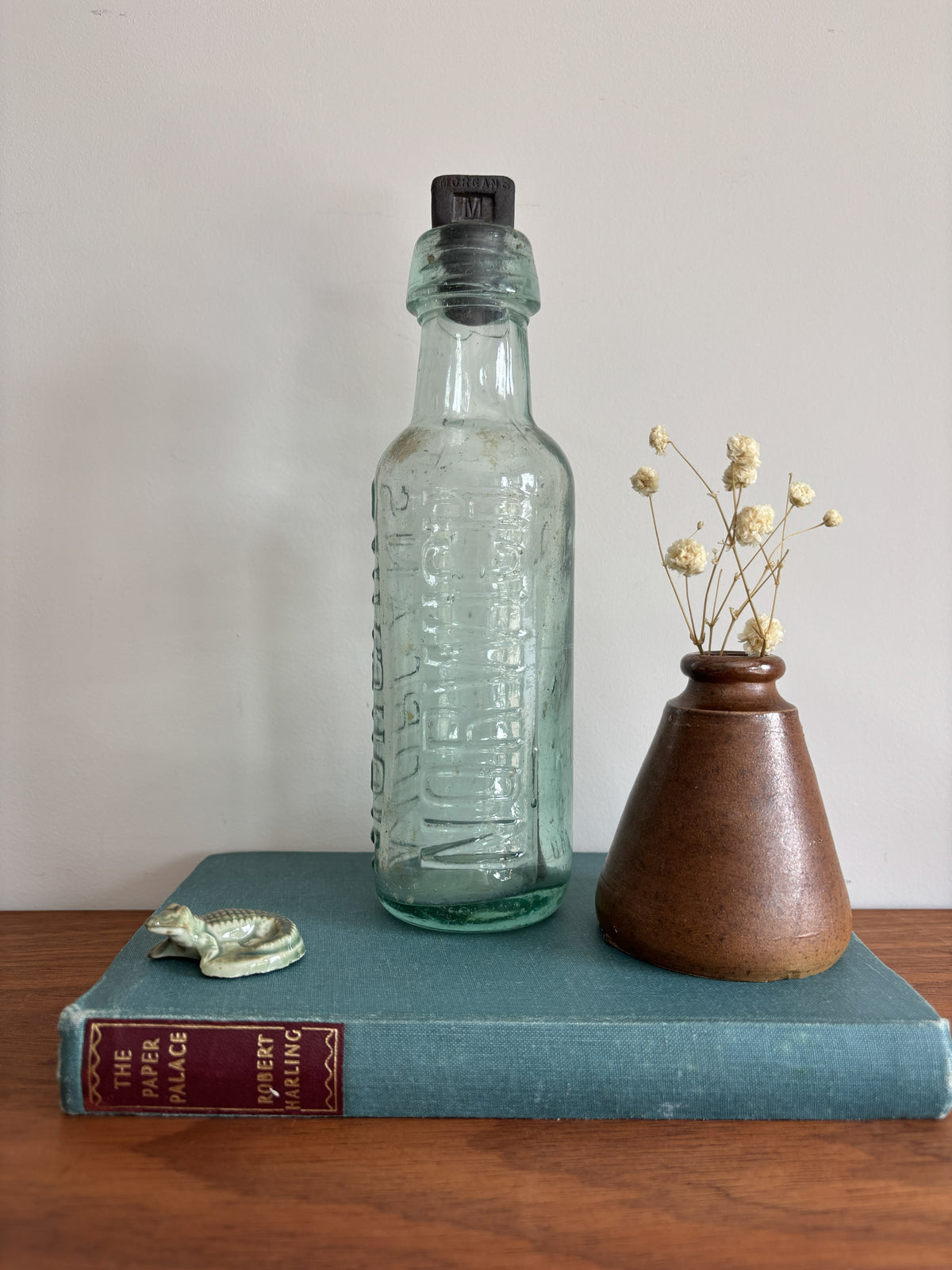 A vintage embossed glass bottle with a black stopper, displayed on a teal book next to a small brown ceramic vase with dried flowers.  The words Morgans and Norwich are embossed on the sides. 