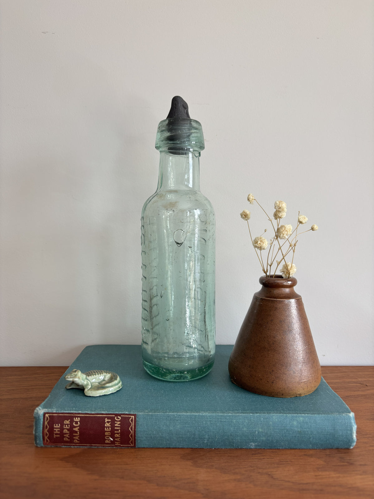 A vintage embossed glass bottle with a black stopper, displayed on a teal book next to a small brown ceramic vase with dried flowers.  The words Morgans and Norwich are embossed on the sides. 