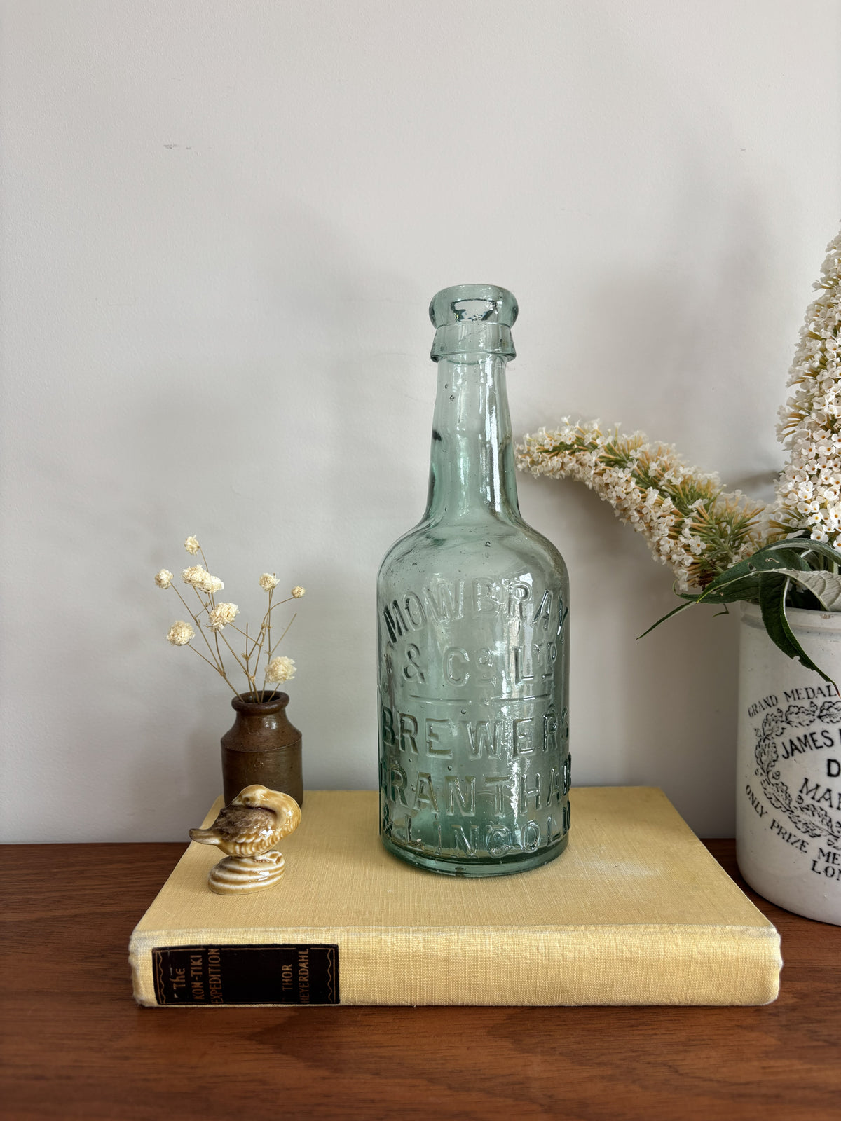 A vintage Mowbray & Co embossed glass bottle in an aqua sea green colour, displayed on a yellow book on a wooden table with decorative dried flowers and a ceramic pot against a plain wall.
