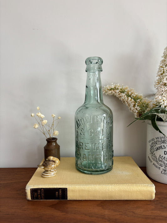 A vintage Mowbray & Co embossed glass bottle in an aqua sea green colour, displayed on a yellow book on a wooden table with decorative dried flowers and a ceramic pot against a plain wall.