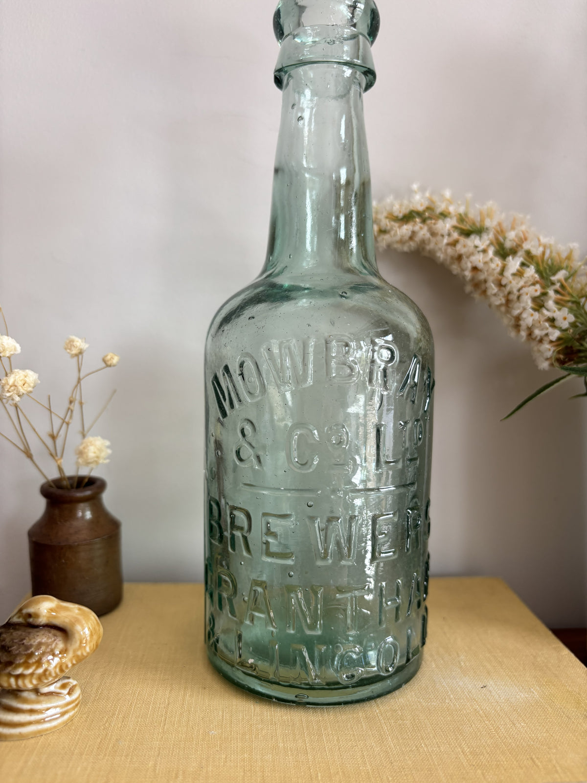 A vintage Mowbray & Co embossed glass bottle in an aqua sea green colour, displayed on a yellow book on a wooden table with decorative dried flowers and a ceramic pot against a plain wall.