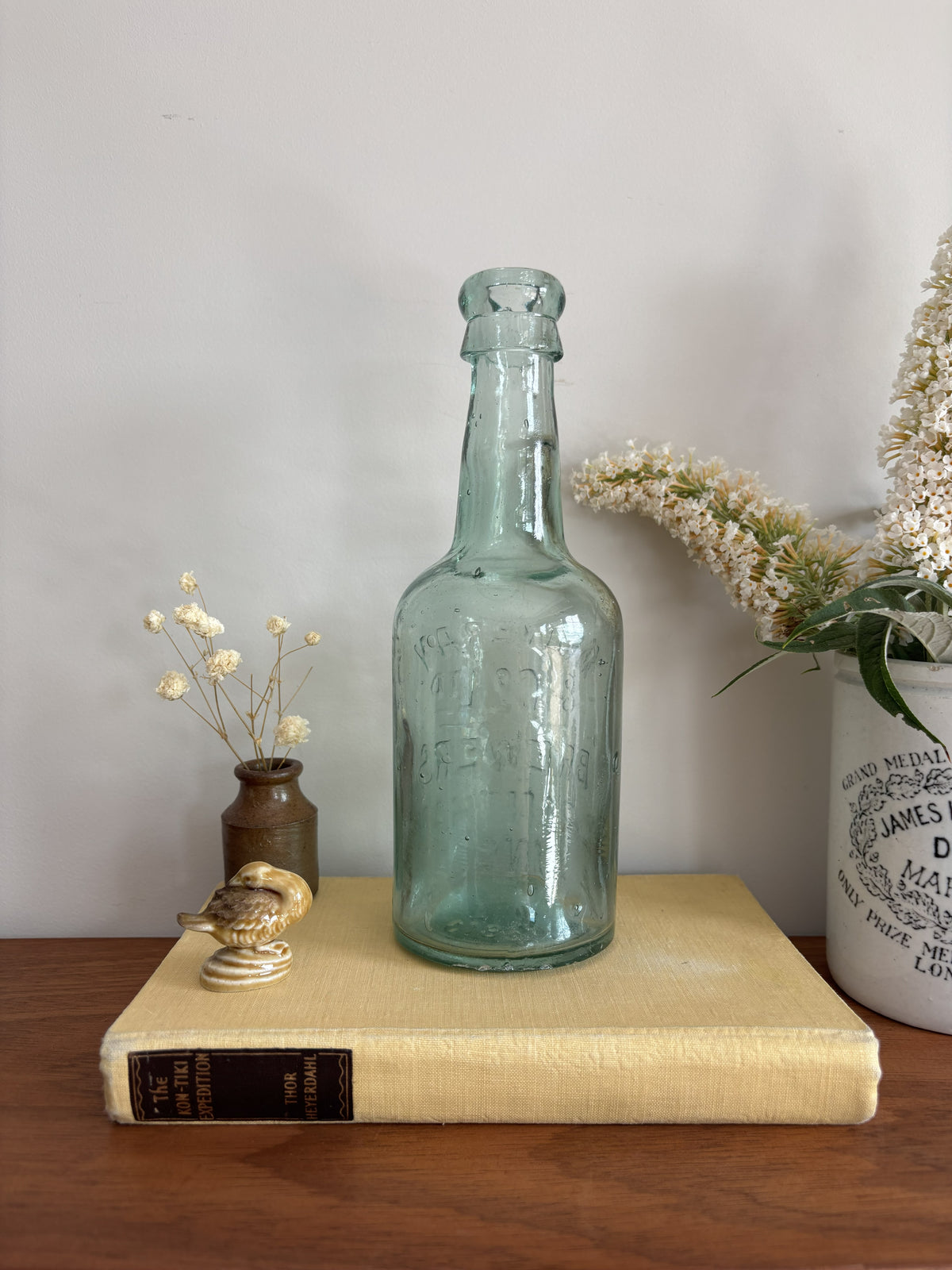 A vintage Mowbray & Co embossed glass bottle in an aqua sea green colour, displayed on a yellow book on a wooden table with decorative dried flowers and a ceramic pot against a plain wall.