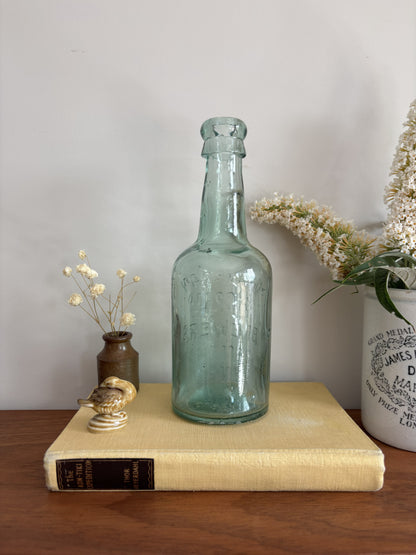 A vintage Mowbray & Co embossed glass bottle in an aqua sea green colour, displayed on a yellow book on a wooden table with decorative dried flowers and a ceramic pot against a plain wall.