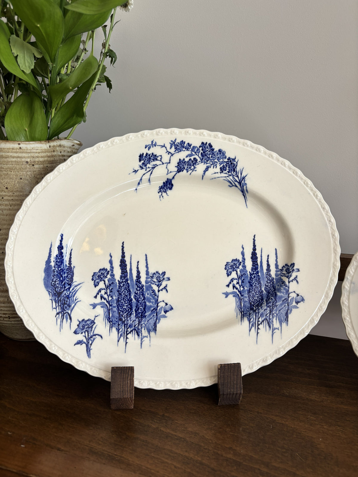 An oval white ceramic serving platter with blue floral patterns displayed on a wooden stand on a dark wood table with a potted plant in the background.
