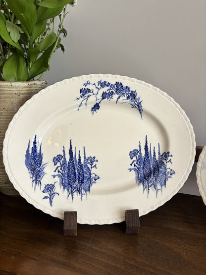 An oval white ceramic serving platter with blue floral patterns displayed on a wooden stand on a dark wood table with a potted plant in the background.