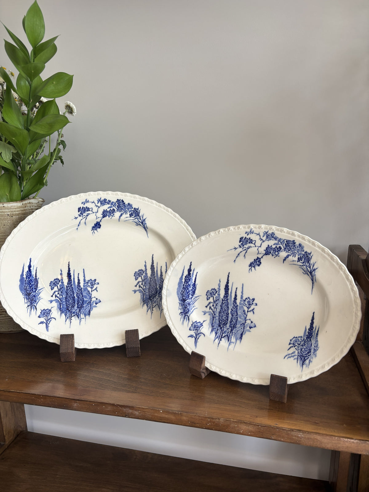 Two oval white ceramic platters with blue floral and botanical illustrations, displayed on small wooden stands on a wooden shelf against a gray wall.