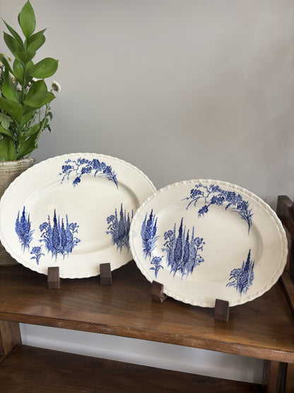 Two oval white ceramic platters with blue floral and botanical illustrations, displayed on small wooden stands on a wooden shelf against a gray wall.