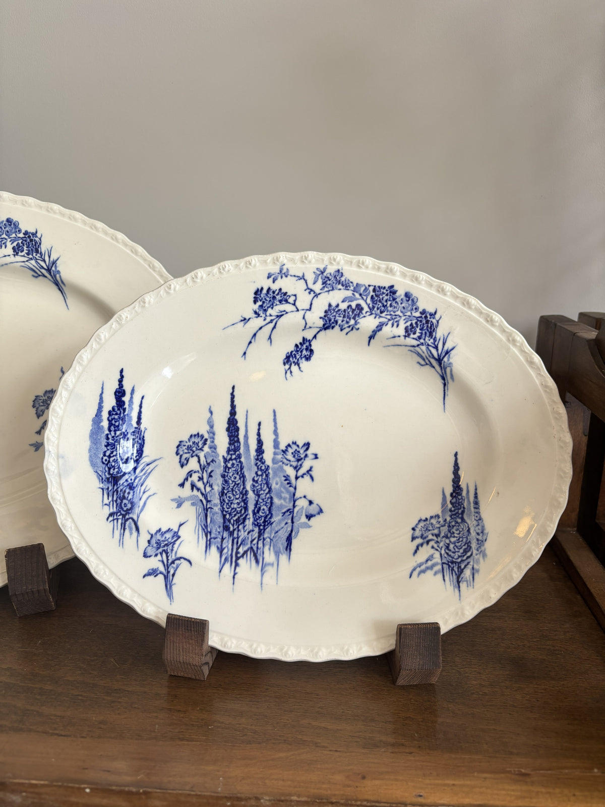 An oval white ceramic serving platter with a blue floral and botanical landscape print, displayed on a wooden stand on a dark wood table.