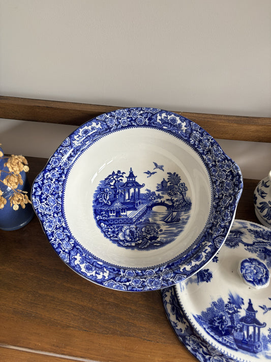 A blue and white tureen dish with a detailed landscape 'willow tree' pattern, displayed on a wooden shelf next to other matching blue and white dishes.