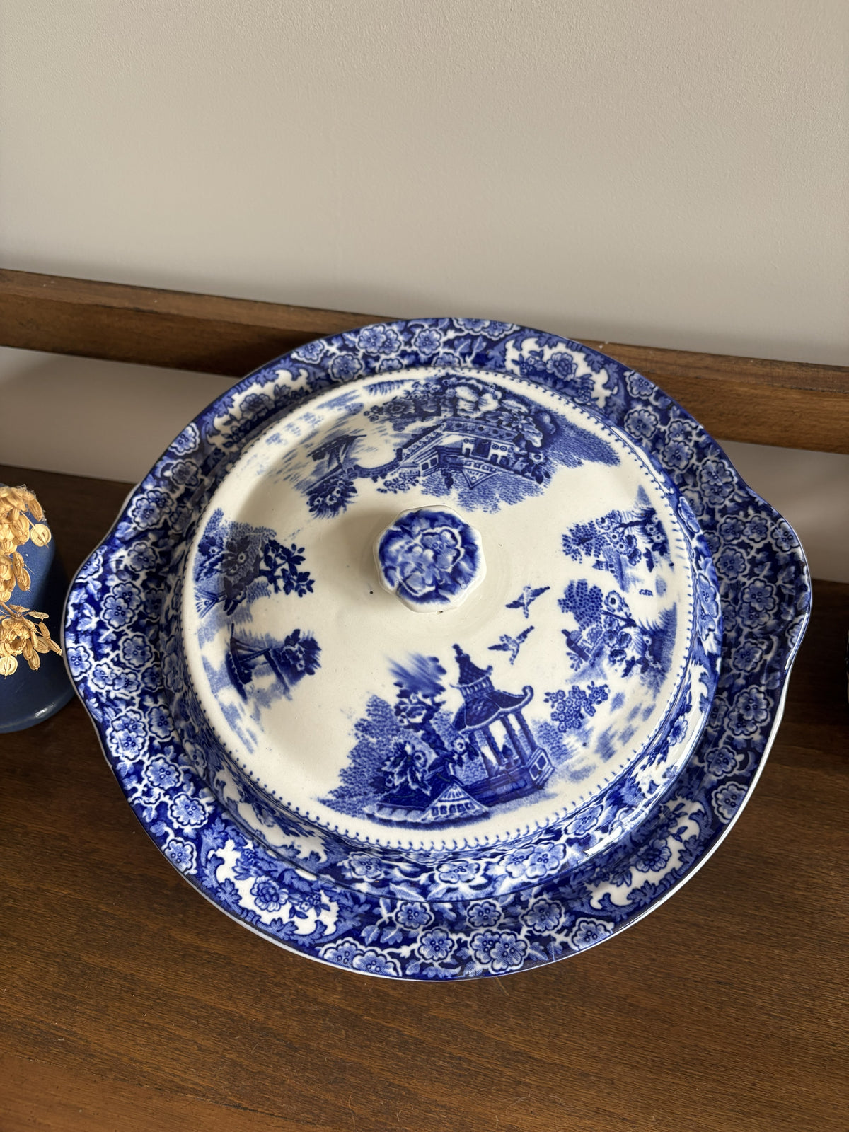 A blue and white ceramic tureen dish with a scenic landscape pattern, resting on a wooden shelf.
