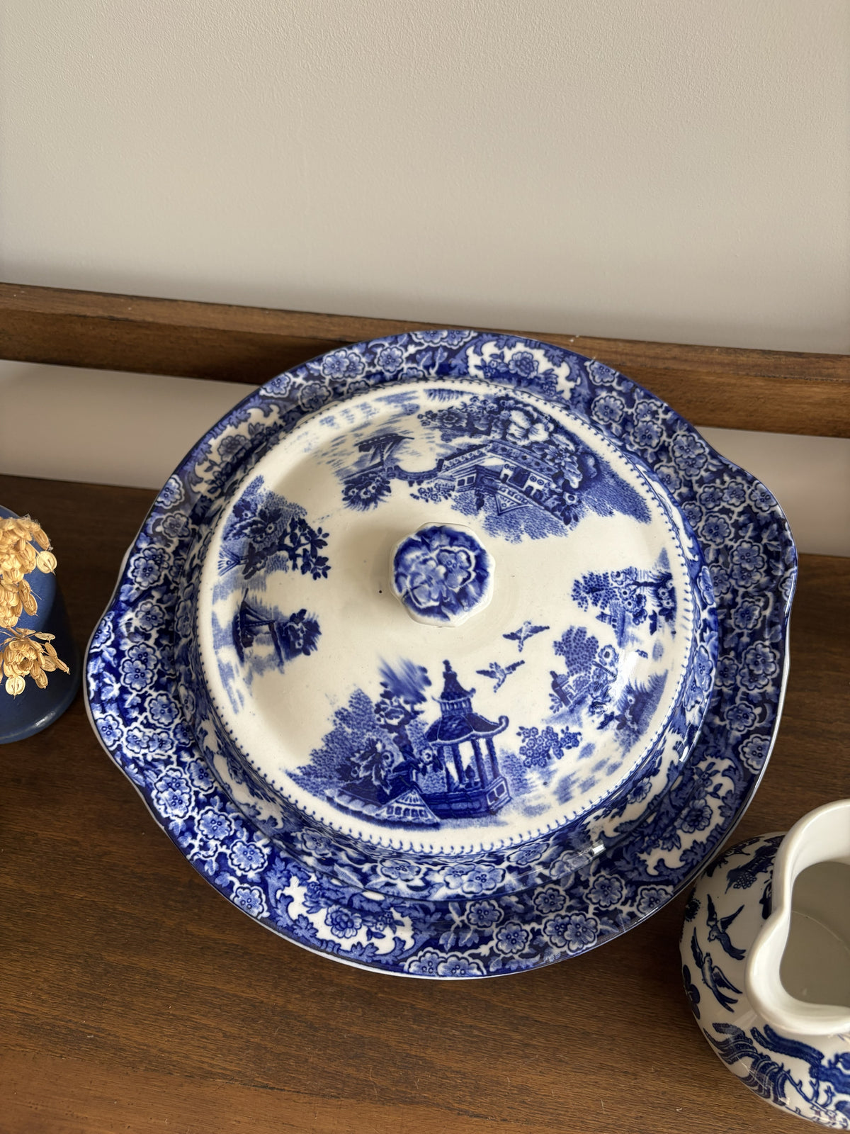 A blue and white lidded tureen dish  with a floral border and scenic landscape illustration, displayed on a wooden surface.