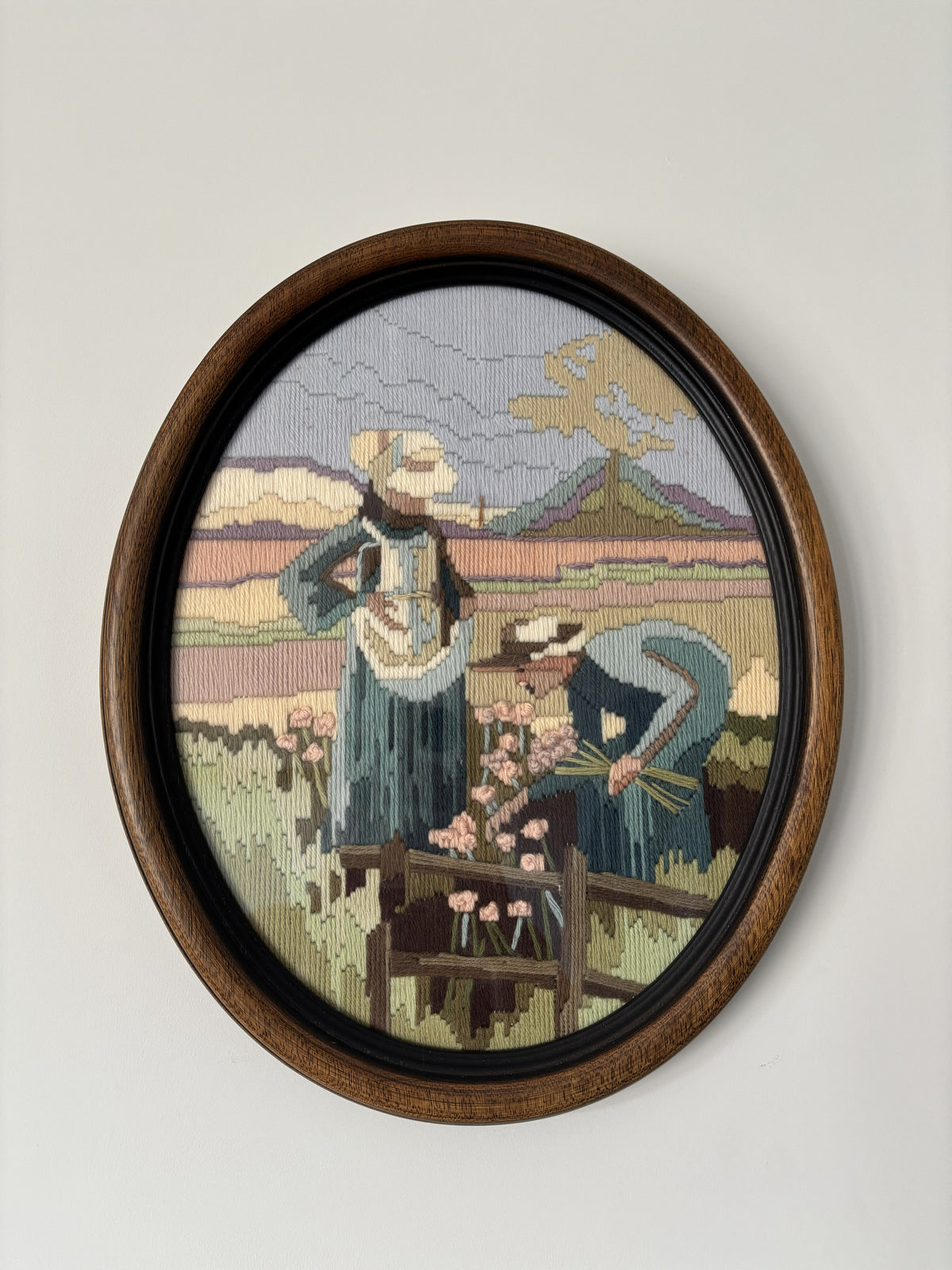 An oval framed needlepoint artwork depicting two women in a field with flowers and mountains in the background, mounted on a light gray wall.