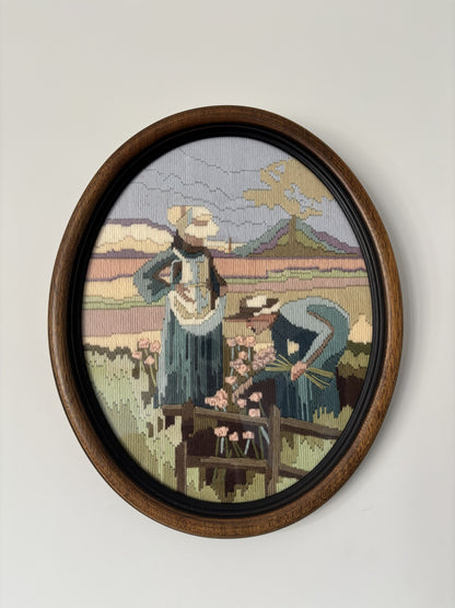 An oval framed needlepoint artwork depicting two women in a field with flowers and mountains in the background, mounted on a light gray wall.