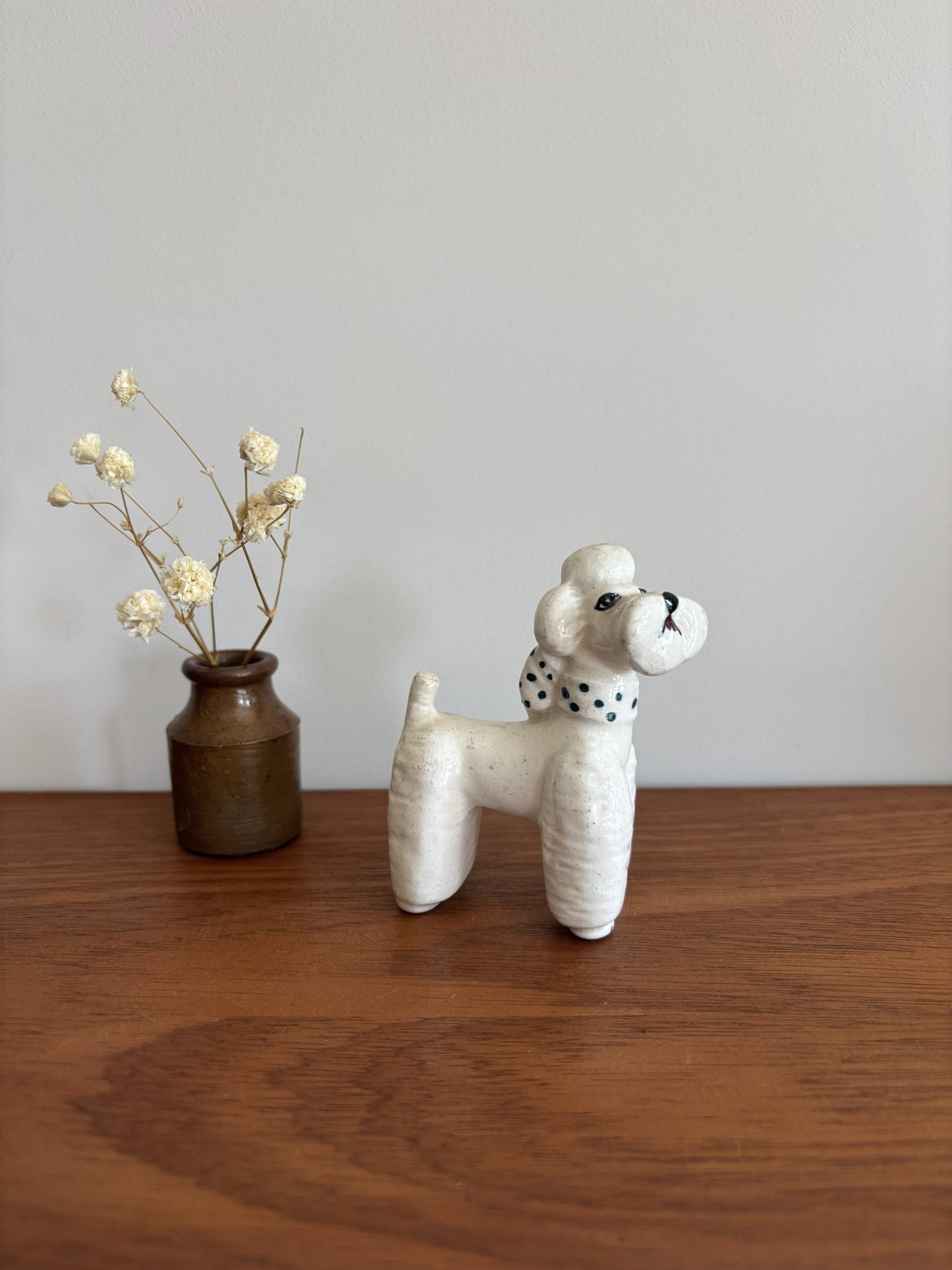 A small white ceramic poodle figurine with a green-and-white polka dot scarf stands on a wooden tabletop against a plain light gray wall. A small brown vase with dried white flowers sits to the left.