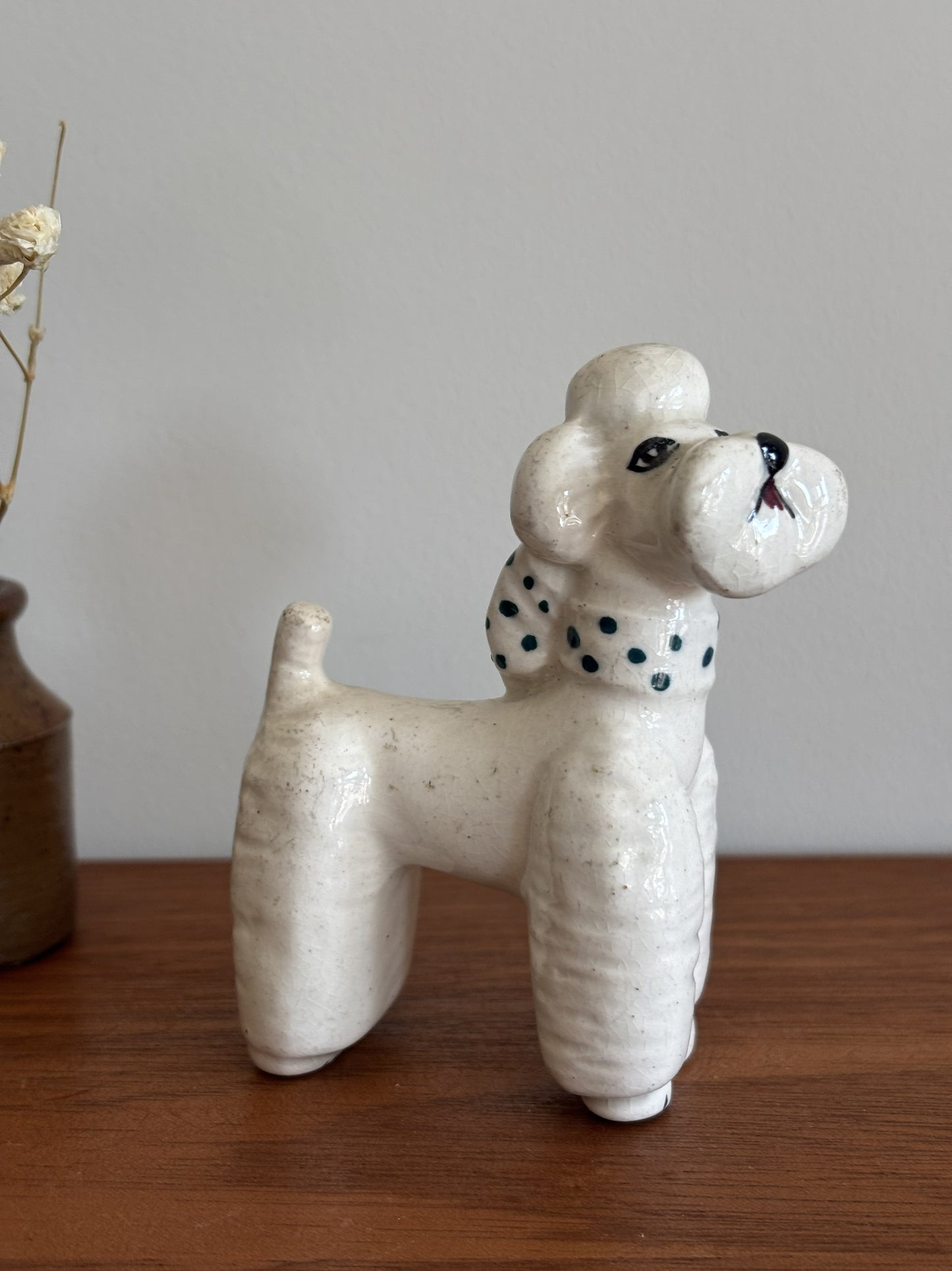 A ceramic poodle figurine in an off-white glaze with a green polka dots on its collar, standing on a wooden surface against a neutral gray background.