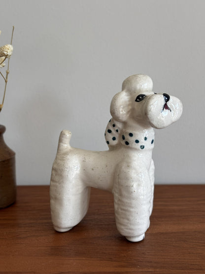 A ceramic poodle figurine in an off-white glaze with a green polka dots on its collar, standing on a wooden surface against a neutral gray background.
