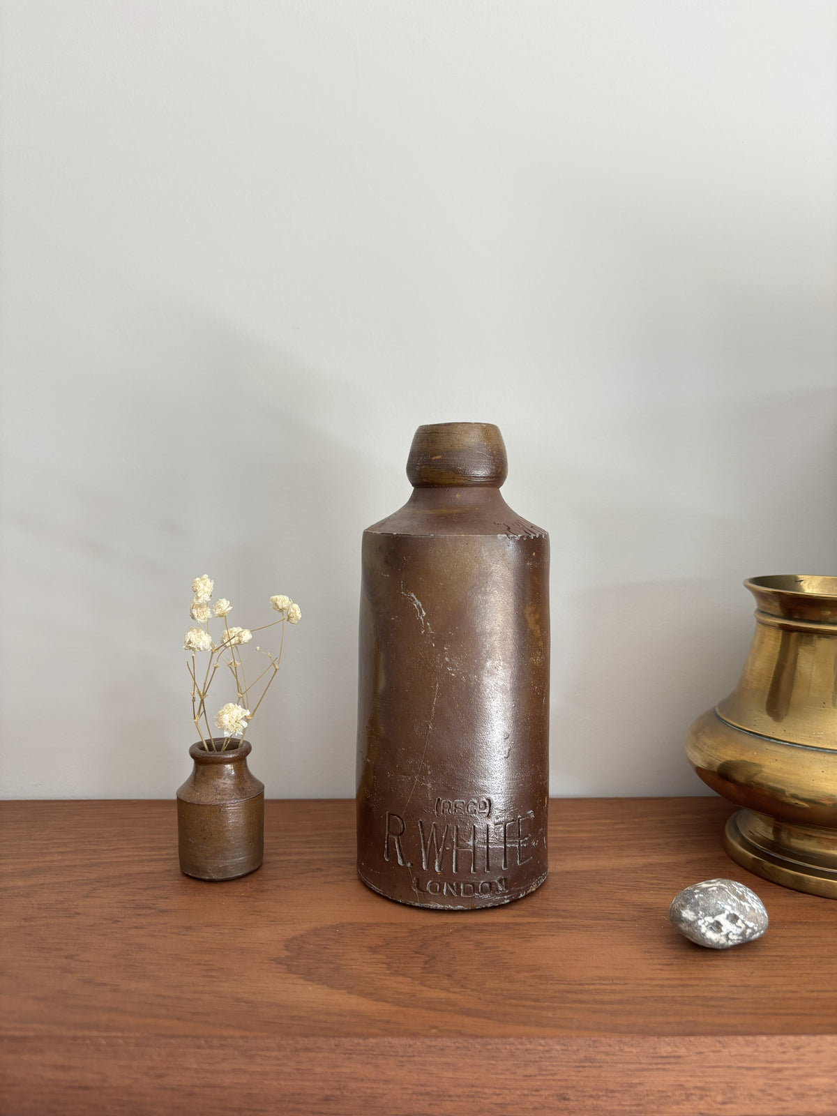 A vintage-brown stone bottle with embossed lettering stands on a wooden surface against a light gray wall, styled with a small vase of dried flowers, a silver stone, and a brass pot.