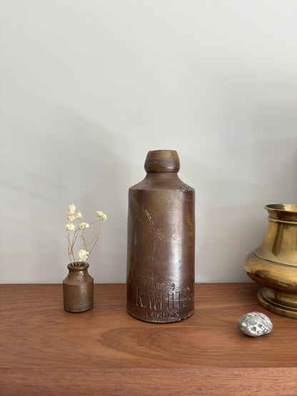 A vintage-brown stone bottle with embossed lettering stands on a wooden surface against a light gray wall, styled with a small vase of dried flowers, a silver stone, and a brass pot.