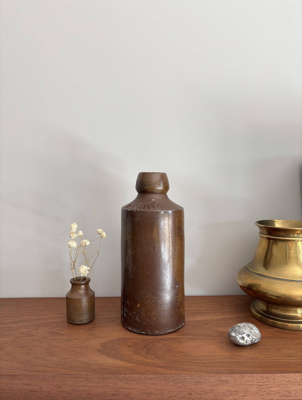 A vintage brown stone bottle, displayed on a wooden surface alongside a small vase with dried flowers, a silver stone, and a brass pot against a plain light gray wall.