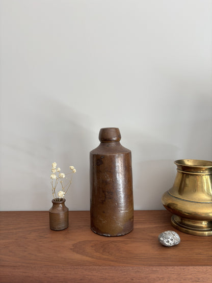 A vintage stone bottle with a rounded neck and flat base, displayed on a wooden surface alongside a small vase with dried flowers, a silver stone, and a brass pot against a plain light gray wall.