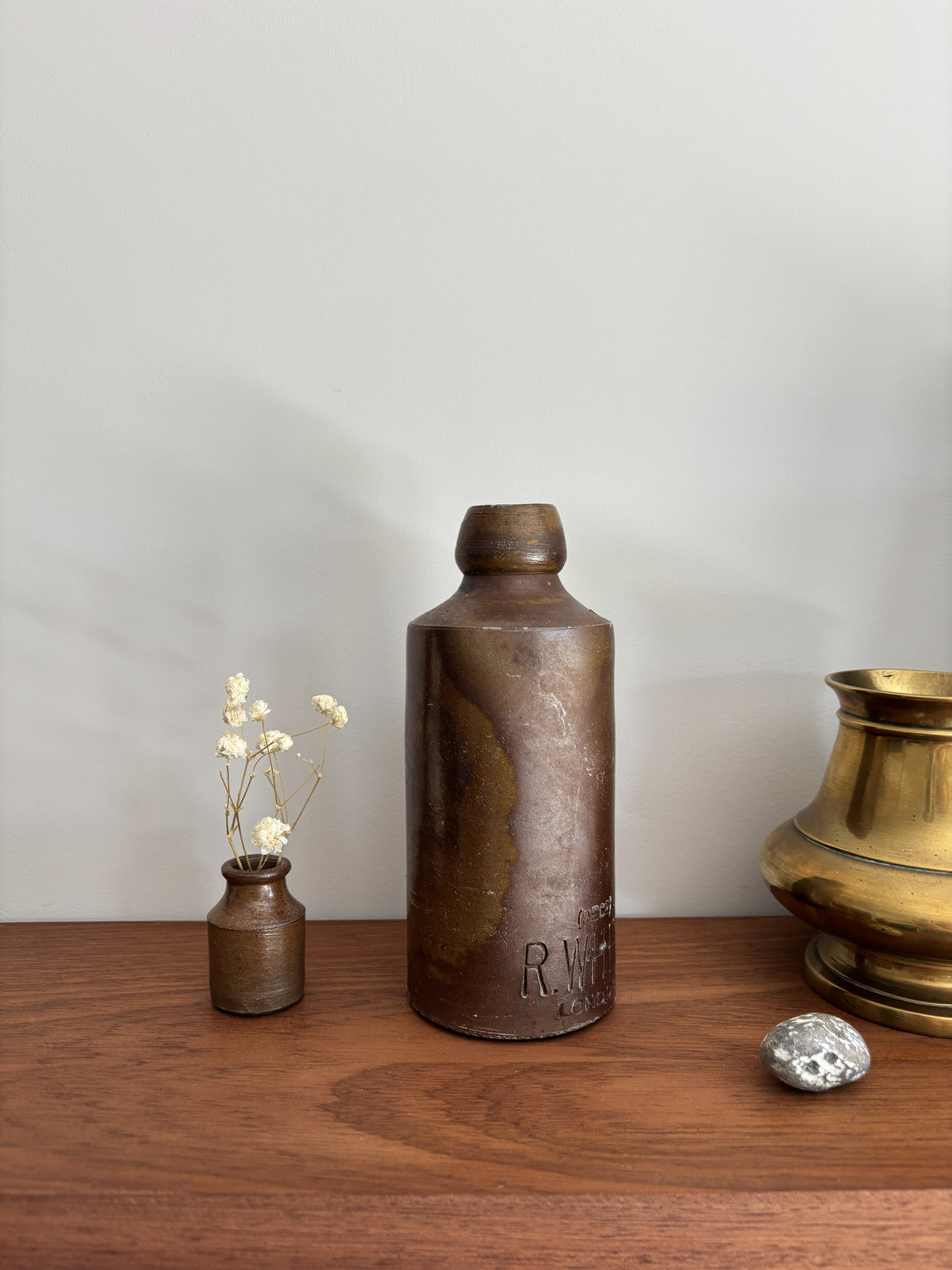 A vintage stone bottle displayed on a wooden shelf alongside a small vase with dried flowers, a polished brass pot, and a small gray stone against a neutral gray wall.