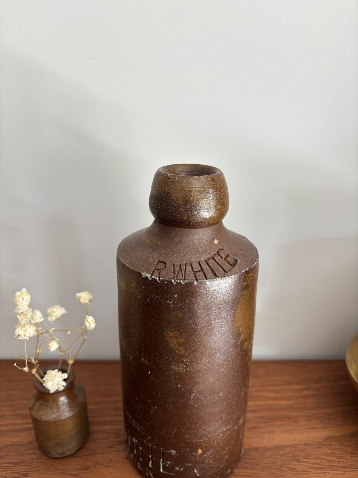 A vintage brown stone bottle with a small neck and embossed lettering, displayed on a wooden surface next to a small vase of dried white flowers against a plain light gray wall.