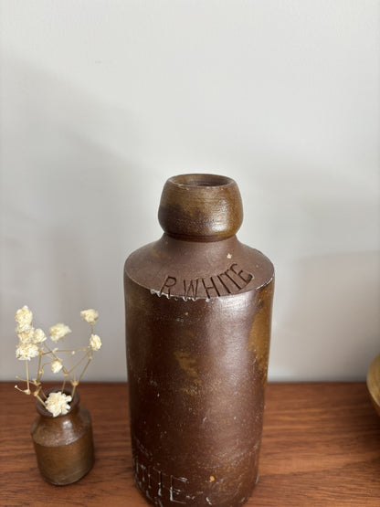 A vintage brown stone bottle with a small neck and embossed lettering, displayed on a wooden surface next to a small vase of dried white flowers against a plain light gray wall.