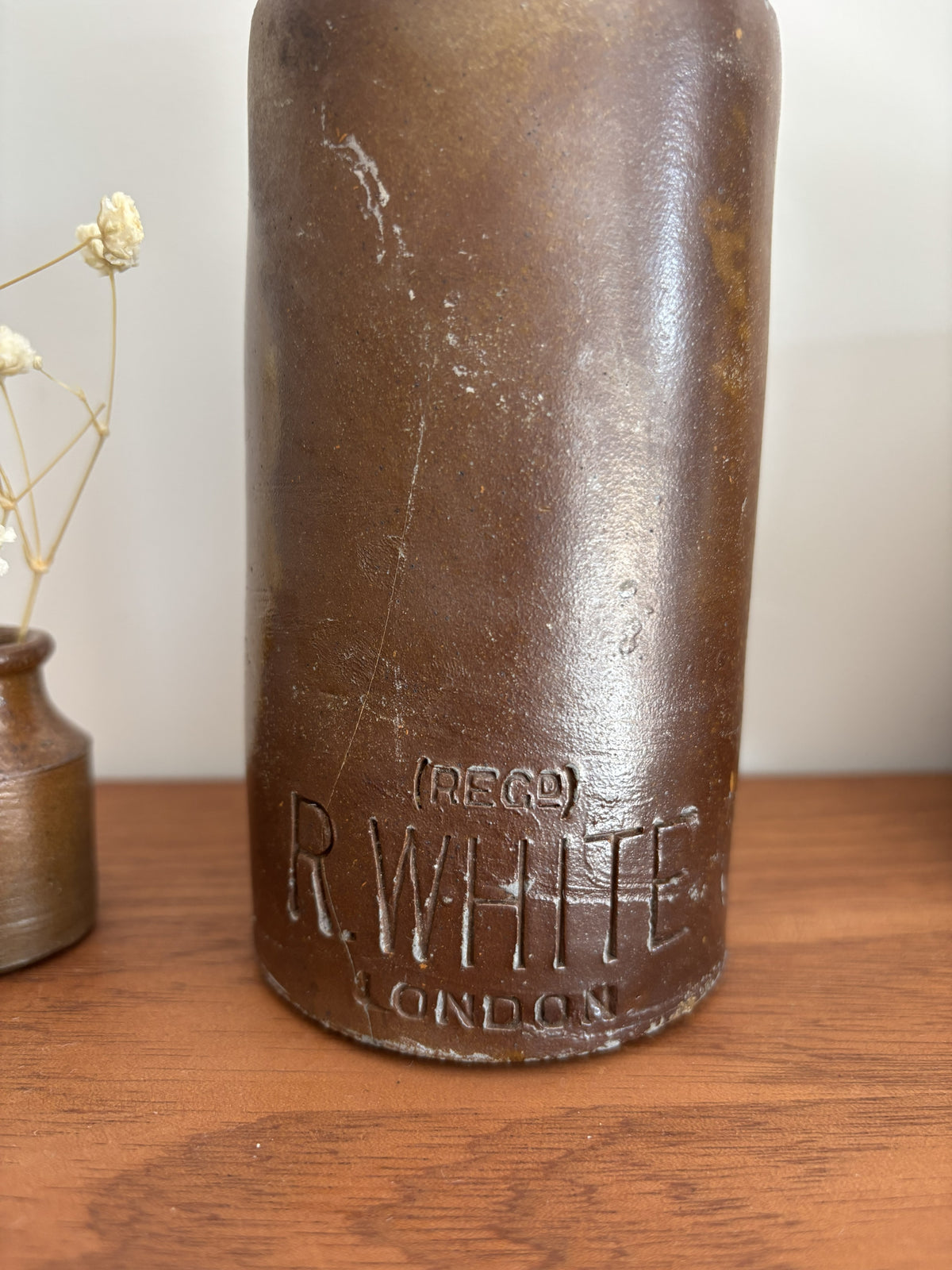 A vintage R. White London stone bottle with embossed lettering, displayed on a wooden tabletop next to a small vase with dried flowers.