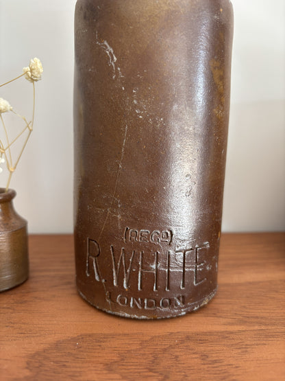 A vintage R. White London stone bottle with embossed lettering, displayed on a wooden tabletop next to a small vase with dried flowers.