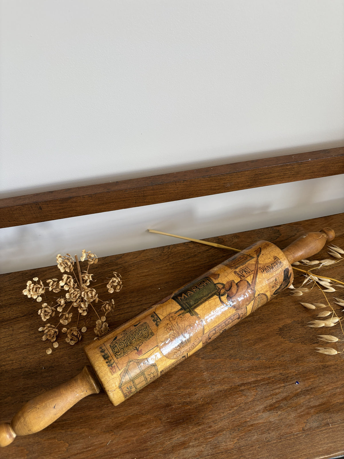 A vintage wooden rolling pin with a printed calendar design rests on a rustic wooden table, styled with dried flowers and wheat stalks against a neutral background.