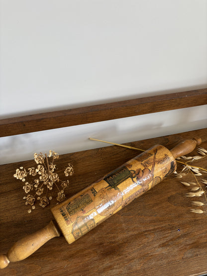 A vintage wooden rolling pin with a printed calendar design rests on a rustic wooden table, styled with dried flowers and wheat stalks against a neutral background.