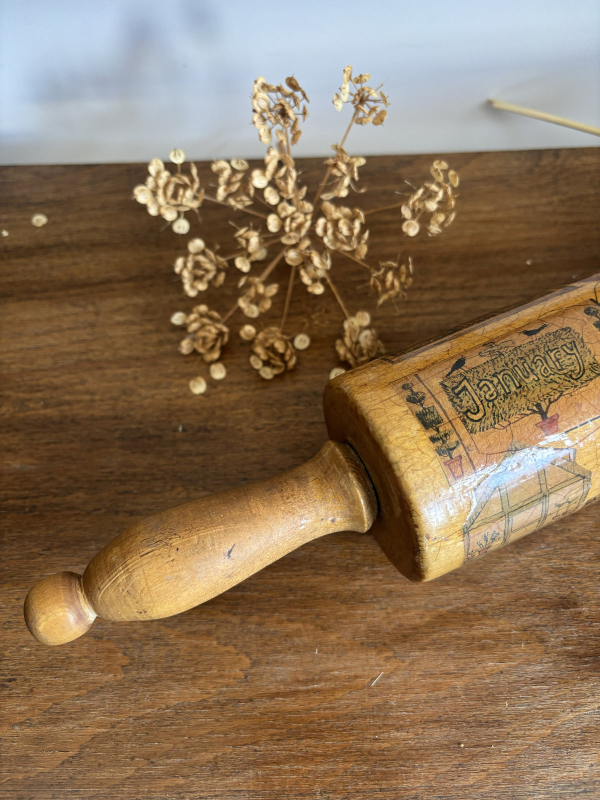 A vintage wooden rolling pin with printed illustrations and text, resting on a rustic wooden tabletop next to dried flowers.