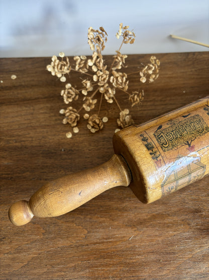 A vintage wooden rolling pin with printed illustrations and text, resting on a rustic wooden tabletop next to dried flowers.