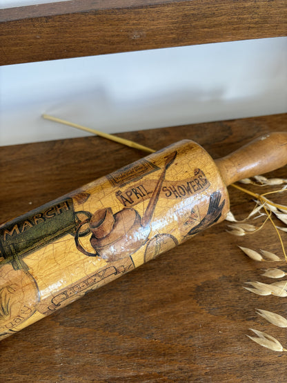 A close-up of a hand-painted wooden rolling pin resting on a rustic wooden surface, decorated with vintage-style weathered lettering and illustrations of rain boots and umbrellas.