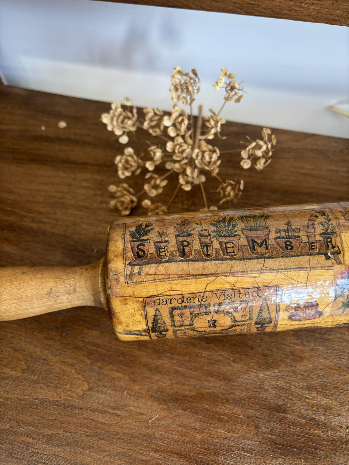 A vintage wooden rolling pin with engraved autumn-themed illustrations and the word 'SEPTEMBER' resting on a wooden tabletop next to dried flowers.