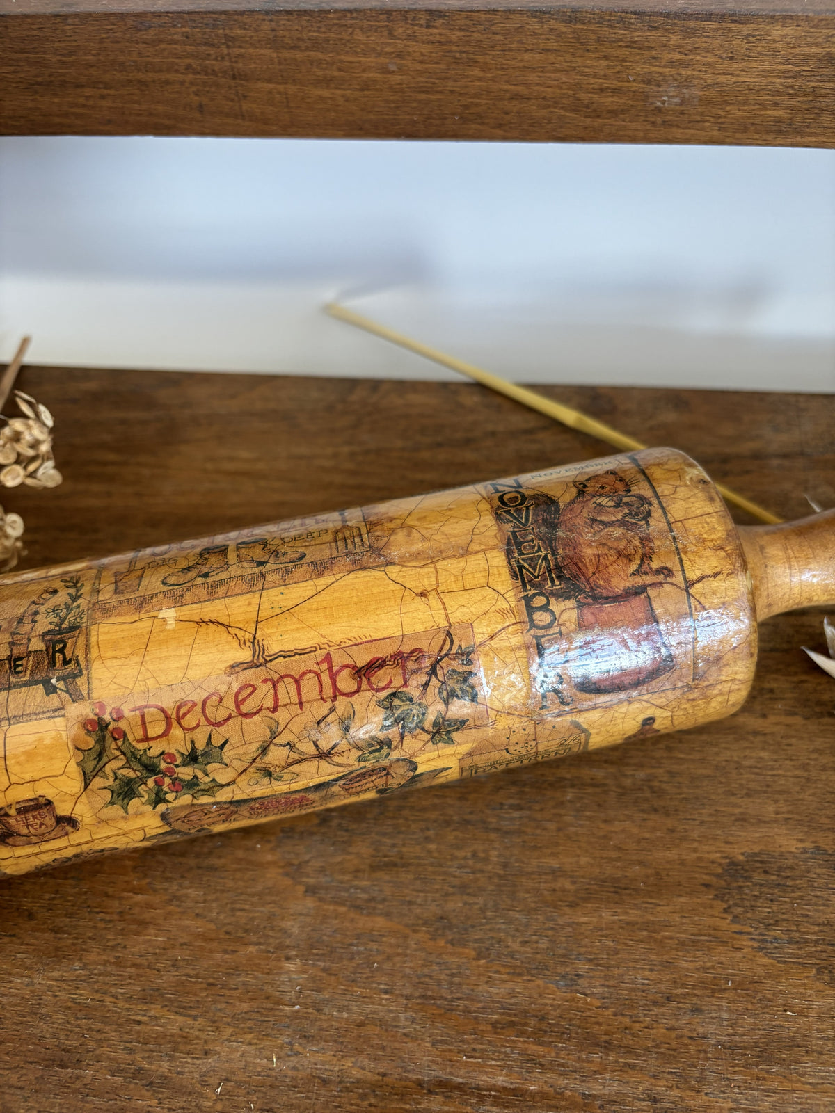 A vintage wooden rolling pin decorated with a detailed Christmas calendar design for the month of December, featuring illustrations and text, resting on a rustic wooden surface.