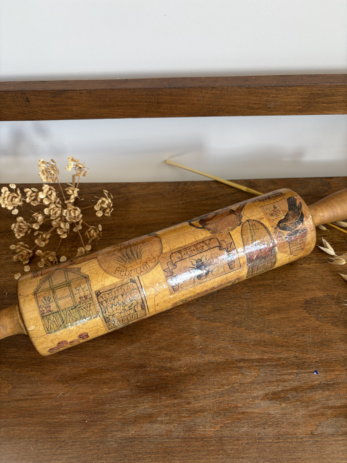 A vintage wooden rolling pin with printed illustrations of houses and scenes, resting on a dark wooden tabletop next to dried flowers.